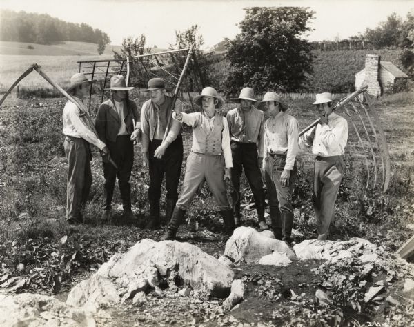 A production still for the Fox Hearst film "Romance of the Reaper". The film was produced by International Harvester at Walnut Grove to celebrate the Reaper Centennial. A group of seven farmers standing, holding scythes and sickles,  with one of the farmers pointing.