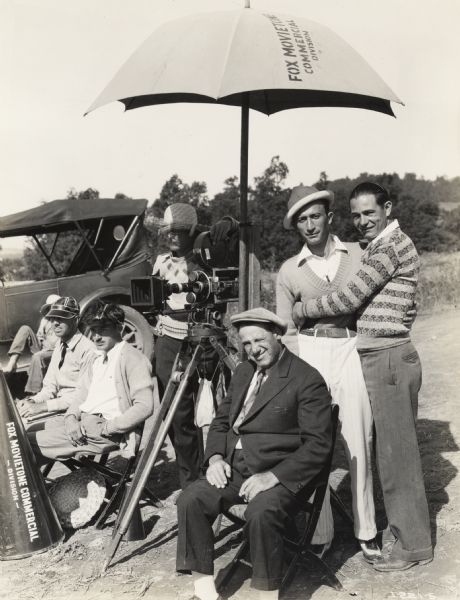 A production still for the Fox Hearst film "Romance of the Reaper." The film was produced by International Harvester at Walnut Grove to celebrate the Reaper Centennial. A group of actors are standing near a cameraman underneath a large umbrella.