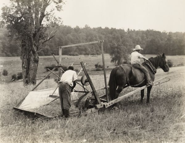 A production still for the Fox Hearst film "Romance of the Reaper". The film was produced by International Harvester at Walnut Grove to celebrate the Reaper Centennial. One man rides a horse which pulls a replica reaper while another man rakes grain.