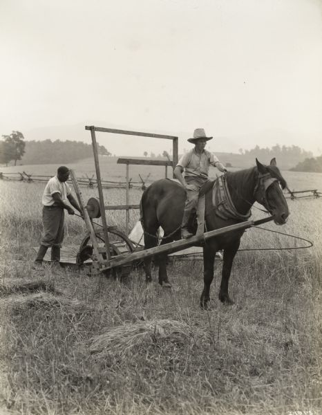 A production still for the Fox Hearst film "Romance of a Reaper". The film was produced by International Harvester at Walnut Grove to celebrate the Reaper Centennial. One man rides a horse which pulls the reaper while another man rakes grain.