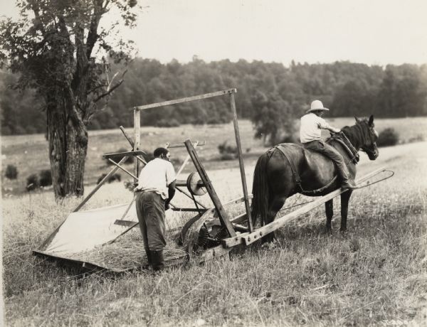 A production still for the Fox Hearst film "Romance of a Reaper". The film was produced by International Harvester at Walnut Grove to celebrate the Reaper Centennial. One man is riding a horse which pulls the reaper while another man is raking grain.