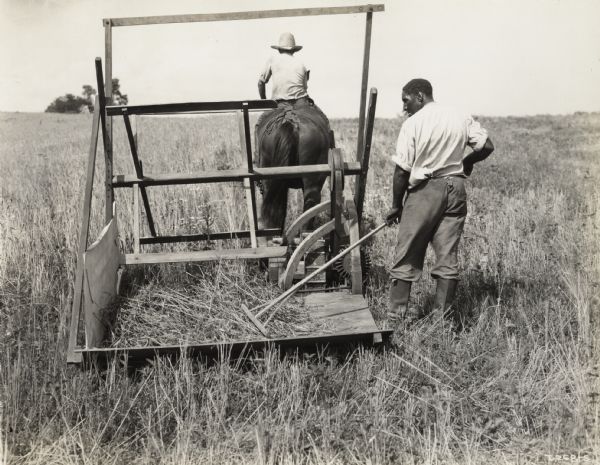 A production still for the Fox Hearst film "Romance of a Reaper". The film was produced by International Harvester at Walnut Grove to celebrate the Reaper Centennial. One man is riding a horse which pulls the reaper, while another man is raking grain.