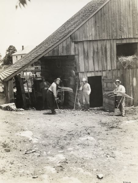 A production still for the Fox Hearst film "Romance of the Reaper". The film was produced by International Harvester at Walnut Grove to celebrate the Reaper Centennial. Four men stand outside of a barn, two of them holding scythes.