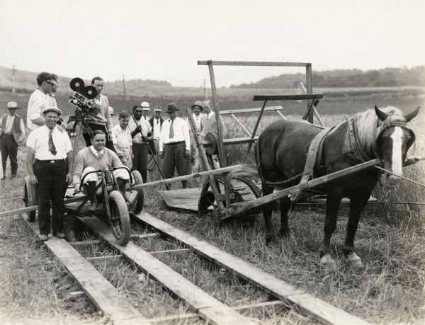 A production still for the Fox Hearst film "Romance of the Reaper". The film was produced by International Harvester at Walnut Grove to celebrate the Reaper Centennial. A group of men are gathered around a camera behind a replica of an 1831 reaper pulled by a horse. The camera is set up on a rolling three-wheeled platform designed to film the reaper in action. The African American man in the center is actor Harry Wilson who played Jo Anderson.