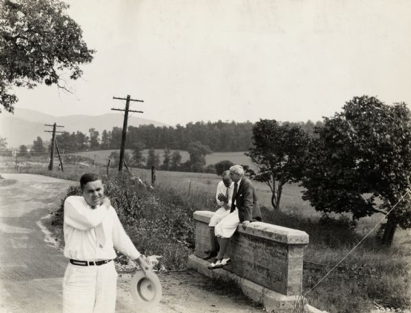 A production still for the Fox Hearst film "Romance of the Reaper." The film was produced by International Harvester at Walnut Grove to celebrate the Reaper Centennial. Two men are resting on a stone ledge, while another man is walking toward the camera in the foreground.