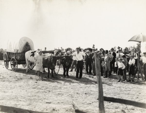 A production still for the Fox Hearst film "Romance of the Reaper". The film was produced by International Harvester at Walnut Grove to celebrate the Reaper Centennial. A large group of men, women, and children stand around a covered wagon pulled by a team of oxen.