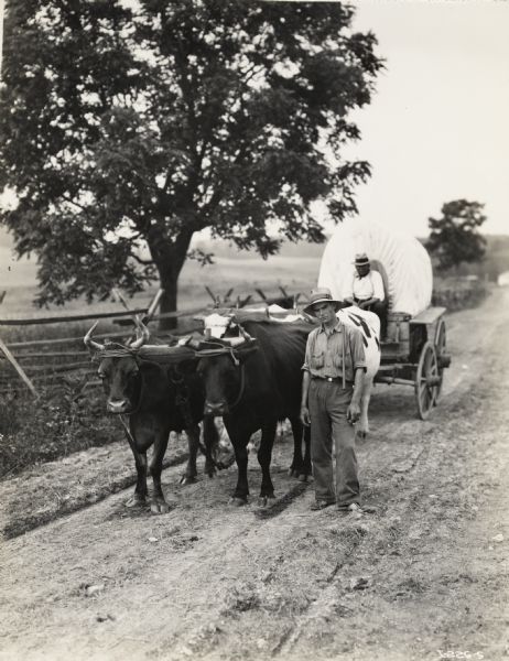 A production still for the Fox Hearst film "Romance of the Reaper." The film was produced by International Harvester at Walnut Grove to celebrate the Reaper Centennial. One man sits on a covered wagon while another stands next to a team of oxen.