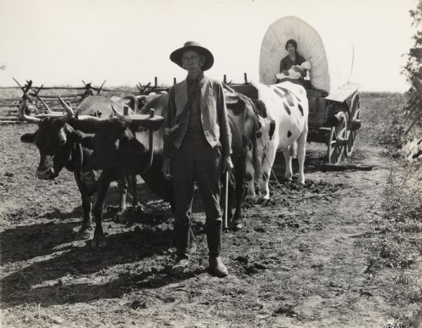 A production still for the Fox Hearst film "Romance of the Reaper." The film was produced by International Harvester at Walnut Grove to celebrate the Reaper Centennial. A man stands in the foreground in front of a team of oxen, and a woman sits on the wagon and holds an infant on her lap.