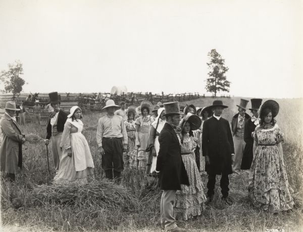 A production still for the Fox Hearst film "Romance of the Reaper." The film was produced by International Harvester at Walnut Grove to celebrate the Reaper Centennial. Actors in actresses in period costume stand in a field on location. In the background are a covered wagon, animals, and other individuals.