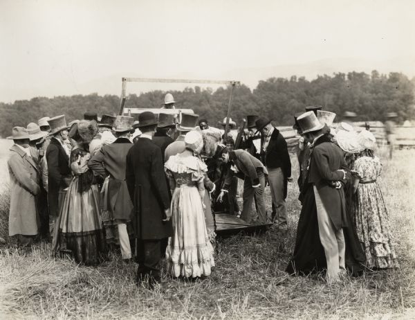A production still for the Fox Hearst film "Romance of the Reaper." The film was produced by International Harvester at Walnut Grove to celebrate the Reaper Centennial. A group of men and woman gather around a replica of a McCormick reaper in a film re-creation of the first reaper test. At the center of the action is actor Del McDermid in the role of Cyrus McCormick.