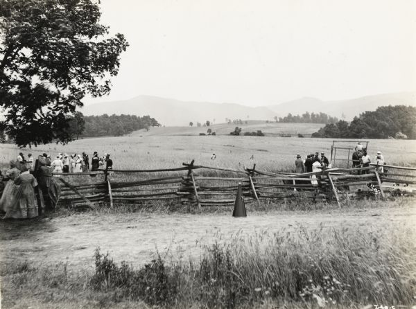 A production still of the Fox Hearst film "Romance of the Reaper." The film was produced by International Harvester at Walnut Grove to celebrate the Reaper Centennial. Several groups of people are gathering near a fence in the foreground. Rolling fields and trees are along the horizon. There is a megaphone sitting on the ground in front of the fence.