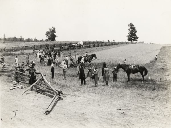 A production still for the Fox Hearst film "Romance of the Reaper." The film was produced by International Harvester at Walnut Grove to celebrate the Reaper Centennial. The cast is gathered in a field for a filmed re-creation of the first reaper test. The actors are arranged to mimic an 1883 advertising lithograph of the reaper test. Del McDermid, in the role of Cyrus McCormick, stands in the foreground holding a hat in his hands, while other men operate a replica reaper in the background.