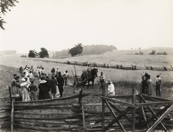 A production still for the Fox Hearst film "Romance of the Reaper." The film was produced by International Harvester at Walnut Grove to celebrate the Reaper Centennial. In this scene men and women are gathered in field to witness the testing of the first McCormick reaper. The reaper used in the film was a replica of McCormick's first reaper.