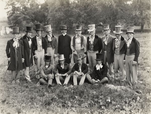 A production still for the Fox Hearst film "Romance of the Reaper." The film was produced by International Harvester at Walnut Grove to celebrate the Reaper Centennial. A group of men in period costume posing in a field on or near location for the film "Romance of the Reaper". Farm buildings are in the background. Many of the actors and extras for the film were local residents.