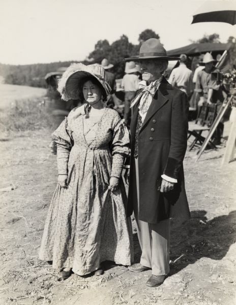 A production still for the Fox Hearst film "Romance of the Reaper." The film was produced by International Harvester at Walnut Grove to celebrate the Reaper Centennial. A woman wearing a period dress and large bonnet stands next to a man wearing a long coat and wide-brimmed hat. The film crew and a camera under an umbrella are in the background.