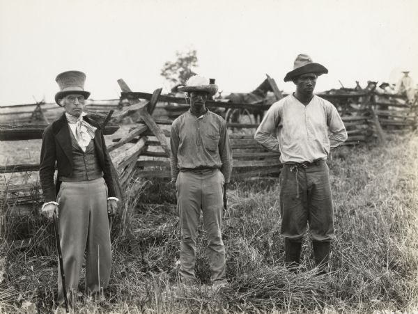 A production still for the Fox Hearst film "Romance of the Reaper." The film was produced by International Harvester at Walnut Grove to celebrate the Reaper Centennial. One well-dressed man with a top hat and cane stands with two men who are dressed as farmers. The men are dressed in period costume and may have been local residents recruited for the film.