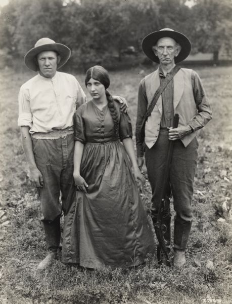 A production still for the Fox Hearst film "Romance of the Reaper." The film was produced by International Harvester at Walnut Grove to celebrate the Reaper Centennial. Two men wearing hats, with one holding a rifle, stand on either side of a woman with a long braid wearing a long dress. The men and woman are dressed in period costume and may have been local residents recruited for the film.