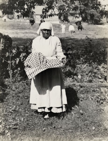 A production still for the Fox Hearst film "Romance of the Reaper". The film was produced by International Harvester at Walnut Grove to celebrate the Reaper Centennial. Clara Wilson, who played the role of a house servant in the film, holds a basket in a field. There is a farm building in the background.