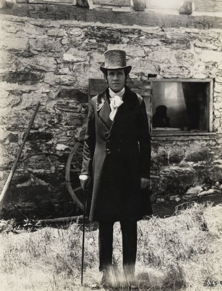 A production still for the Fox Hearst film "Romance of the Reaper". The film was produced by International Harvester at Walnut Grove to celebrate the Reaper Centennial. A man wearing a top hat and suit, and holding a cane is standing in front of the McCormick blacksmith shop. The man may have been a local resident recruited for the film. A silhouette of another man can be seen inside the window of the building.
