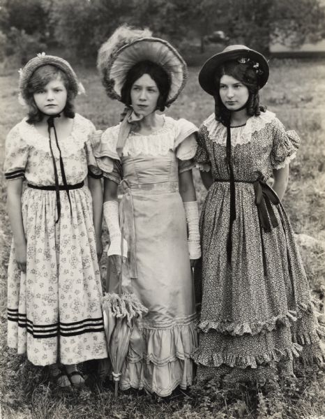 A production still for the Fox Hearst film "Romance of the Reaper". The film was produced by International Harvester at Walnut Grove to celebrate the Reaper Centennial. Three well-dressed, young women are standing in a field. The women may have been local residents recruited for the film.