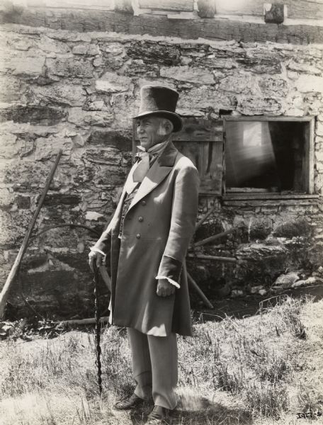A production still for the Fox Hearst film "Romance of the Reaper". The film was produced by International Harvester at Walnut Grove to celebrate the Reaper Centennial. An elderly man wearing a top hat and suit, and holding a cane is standing in front of the McCormick blacksmith shop. The man may have been a local resident recruited for the film.