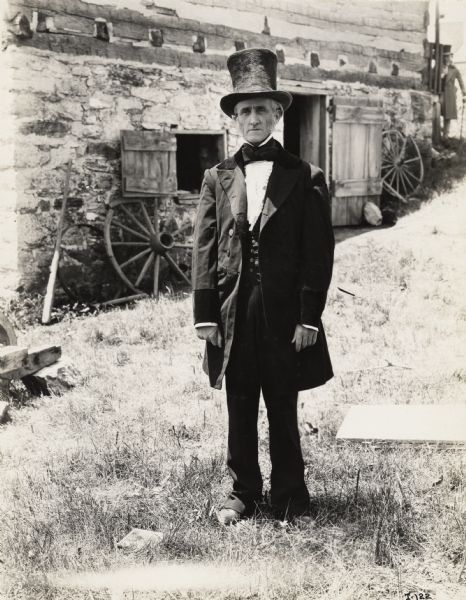 A production still for the Fox Hearst film "Romance of the Reaper". The film was produced by International Harvester at Walnut Grove to celebrate the Reaper Centennial. An elderly man wearing a top hat and suit is standing in front of the McCormick blacksmith shop. The man may have been a local resident recruited for the film.