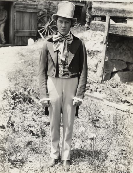 A production still for the Fox Hearst film "Romance of the Reaper." The film was produced by International Harvester at Walnut Grove to celebrate the Reaper Centennial. A young man wearing a light-colored top hat and suit is standing outside of the McCormick blacksmith shop. The man may have been a local resident recruited for the film.