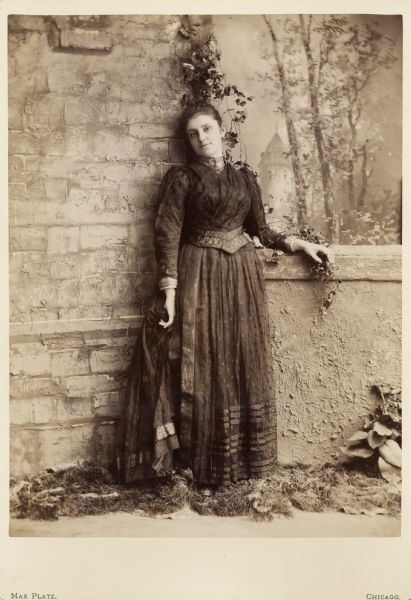 Studio portrait of Mary Virginia McCormick (1861-1941). Mary Virginia was the daughter of Chicago industrialist and inventor, Cyrus Hall McCormick.  She is wearing a long gown and is standing beside a "wall" in front of a painted backdrop.