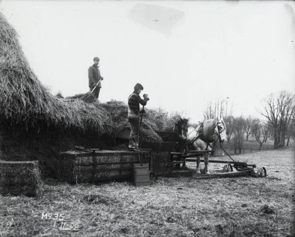 Two men operating a hay press in a mown field. Two horses are hitched to the machine. 
