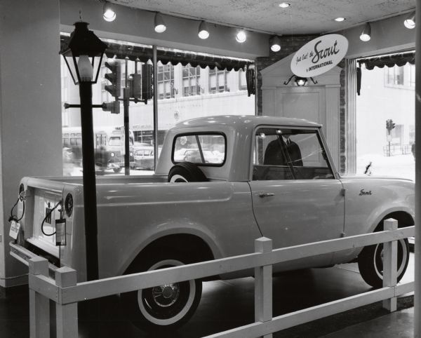 A International Scout truck on display in a showroom. A man is standing behind the truck, which is displayed in front of large windows which look out onto the intersection of a busy city street. This display may have been at the International Harvester headquarters in Chicago, Illinois.