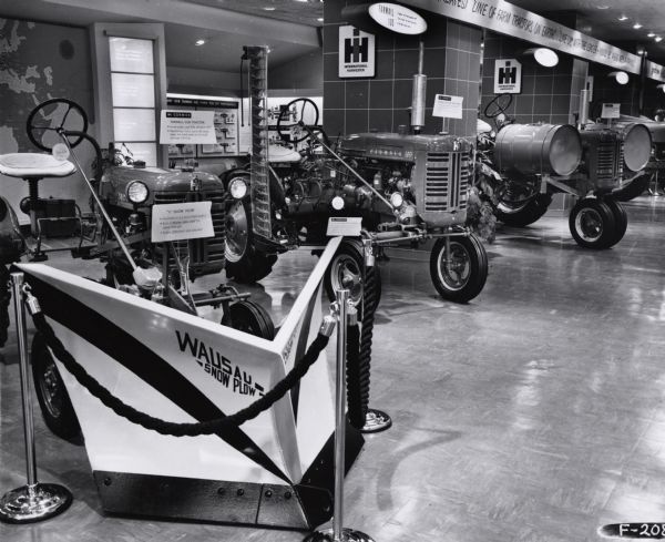 Several tractors are on display in a showroom along with a snowplow. On display are the "V" Wausau Snow Plow, Farmall Cub Tractor, A-22 Side-Mounted Mower and a Farmall 100 Fast-Hitch. The showroom may have been at the International Harvester headquarters building in Chicago, Illinois.