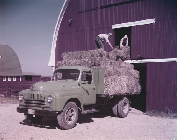 View of an International L-160 Truck with platform body. Men are loading bales of hay from the back of the truck into a barn.