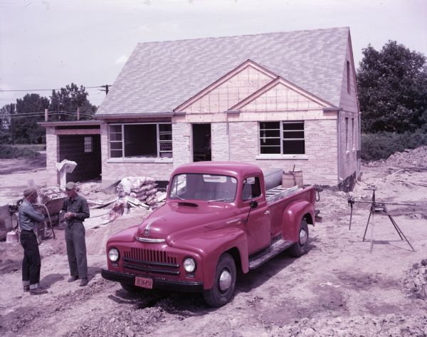View of an International L-120 truck with pickup body parked at a residential construction site. Two men in work clothes and hats stand near the truck.