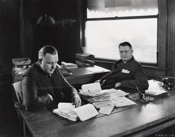 Two men sit at a large desk and look over paperwork. The original caption reads: "E.W. and W.L. Murphy [of] Gateway City Transfer Co. Inc. 'Automotive Admirals' for June 1936 Trail. Taken by Madison Branch." Gateway City Transfer Co. used International trucks.
