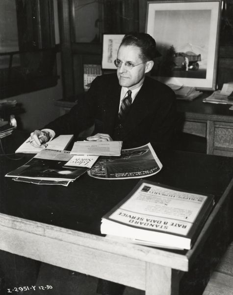 A man sits at a desk and holds a pen in his hand as he looks over promotional paperwork. Caption on photograph reads: "W.L. Nenobuck, Advg. Dept."