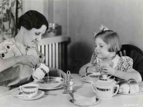 A woman pouring milk or cream into bowl as a young girl, who is holding a glass of milk, is watching.