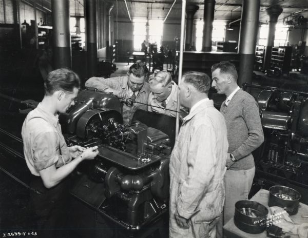 Five men gather around an engine in a factory (most likely International Harvester's Milwaukee Works). Original caption reads: "Branch service managers view a machining operation. Taken by McQuinn".