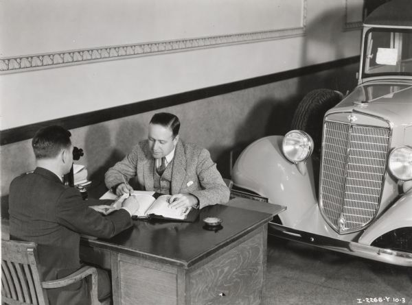 Two men sit at a desk near an International station wagon (possibly in an International truck dealership). Original caption reads: "City Sales, Dan Strong and Bill Koon and Station Wagon. Taken by B.B. Hyde."