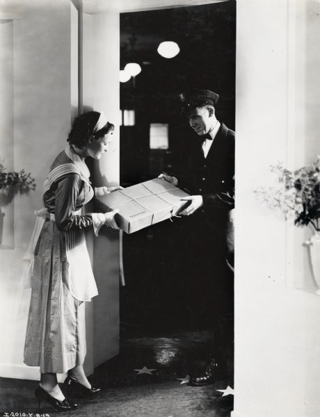 A maid(?) is receiving a package from a delivery man. Original caption  reads: "Posed for Mandel Bros. Story. Taken by K and F." The image was likely used for a story in an "International Harvester" magazine.