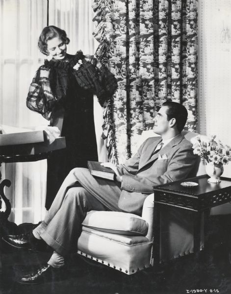 A woman is sstanding and showing a dress to a man who is sitting in a chair. Caption on photograph reads: "Posed for Mandel Bros. Story. Taken by K and F." The image was likely used for a story in an "International Harvester" magazine.