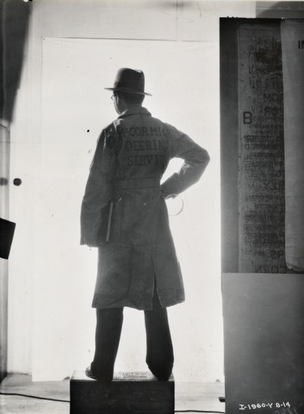 A man stands on a wood box in front of a brightly lit background with his back to the camera. On the back of his coat is written: "McCormick-Deering Service."