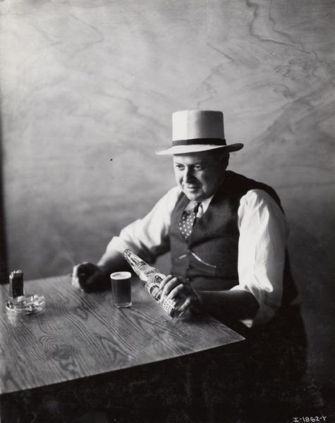 A man is sitting at a table holding a cigarette in one hand and pouring a bottle of Tivoli pilsner beer into a glass with the other. Original caption reads: "Special photos taken for Commercial Car Journal to illustrate article on Tivoli Brewery."