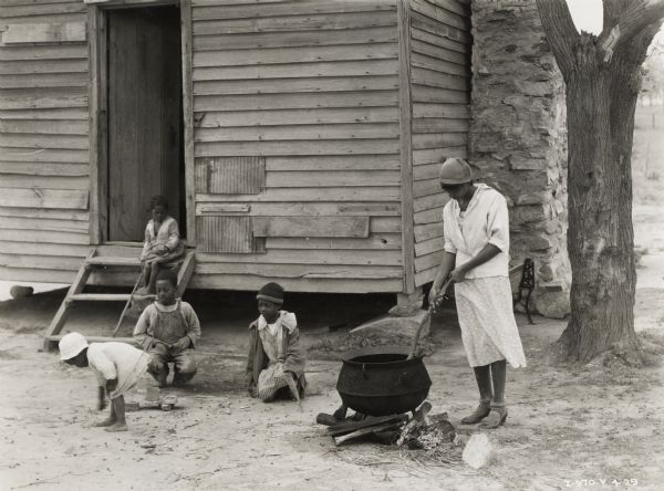 A woman outdoors next to a house standing over a large, black pot, possibly doing laundry. Four children are playing around the woman near the entrance.