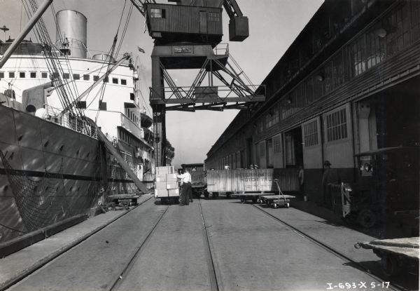 Men loading large "International Motor Truck" crates onto a ship with a crane. Men are working near the ship, and people are watching from the second-story of the building on the right.