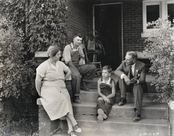 Mr. and Mrs. C.P. Mills and their son Charles of Route 3, Homer, Illinois. A second man, identified in the original caption as "H.G. Davis, N.A.F.E.M." sits with the family on the porch of their home.