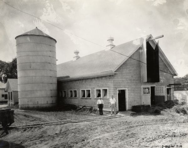 Two men stand outside a barn, with a silo nearby.