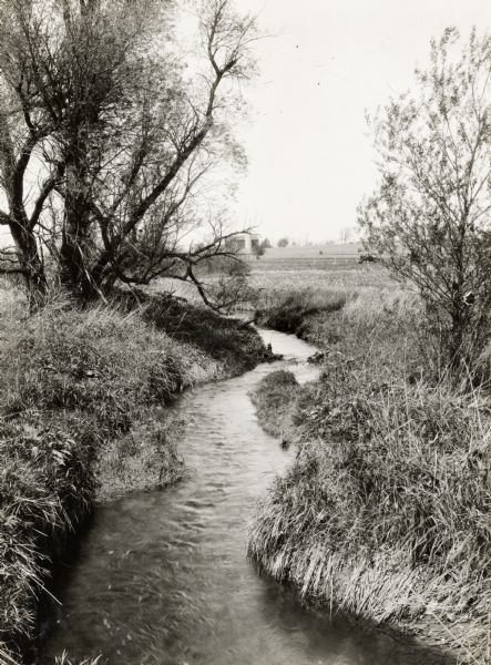 A landscape view of a winding stream and surrounding croplands. A barn and silo are in the distance.