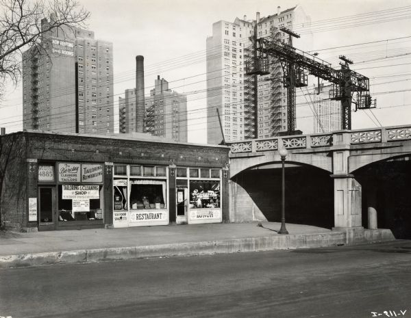 A "Tailoring and Remodeling" shop and restaurant are in the foreground at the entrance to a bridge, while a large utility pole with power lines, a number of tall apartment buildings, and a smokestack are in the background. The bridge may be part of Chicago's elevated train system.