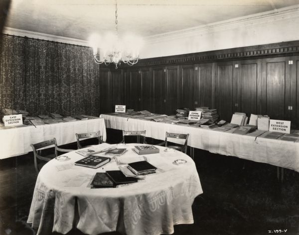 Room with six tables containing what appears to be an informational display of literature created by International Harvester's Agricultural Extension Department. Signs with the display read: "East Extension Section, South Extension Section, Central Extension Section" and "West Extension Section."