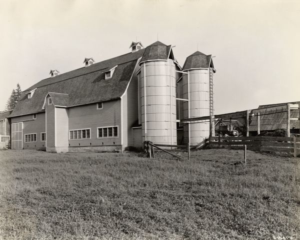Exterior view of a barn and its two attached silos. Adjacent to the barn there is a fenced-in area with two horses and a rider wearing a hat. Farm located outside Cloverdale, British Columbia, Canada.